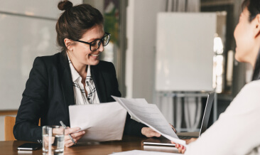 Two business women smiling and talking while looking at papers.